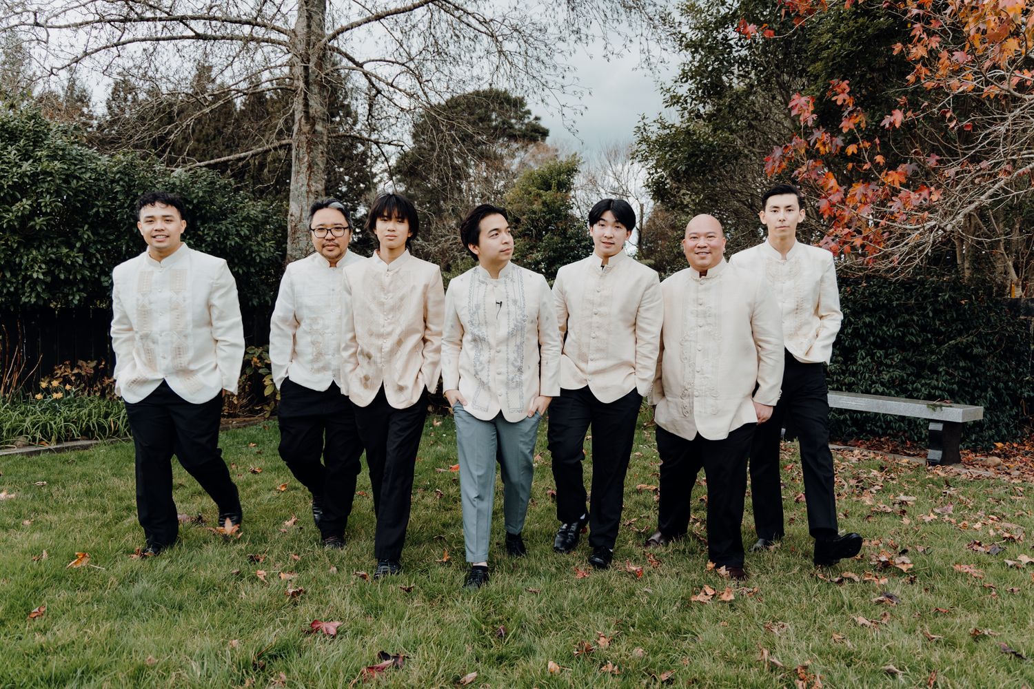 Seven people in coordinated white formal wear stand together in a fall garden setting.