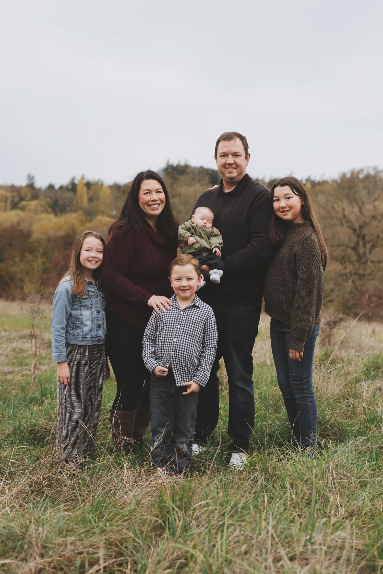 A family of six poses together outdoors in casual fall attire against an autumn landscape with tall grass and trees.