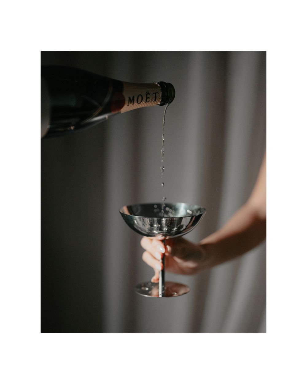 Champagne pouring from bottle into a metallic coupe glass in a moody, dim setting.