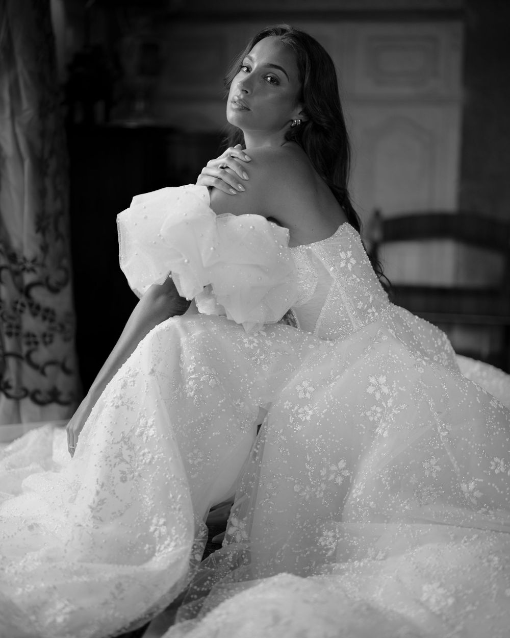 A person in a ruffled white dress poses elegantly in a black and white photograph with dramatic lighting.