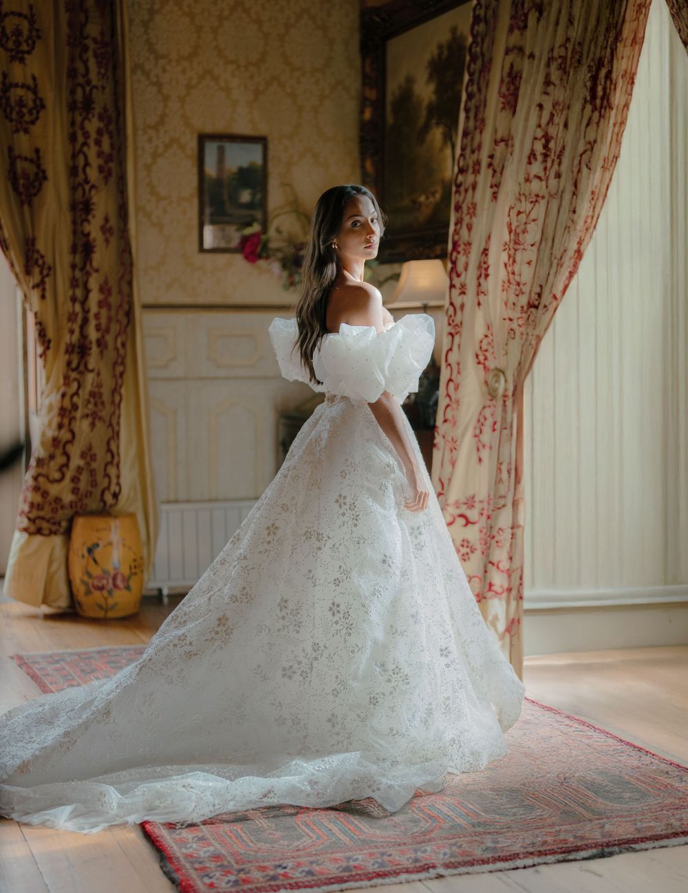 A bridal gown with intricate lace detailing and flowing train spreads elegantly across a patterned red carpet.