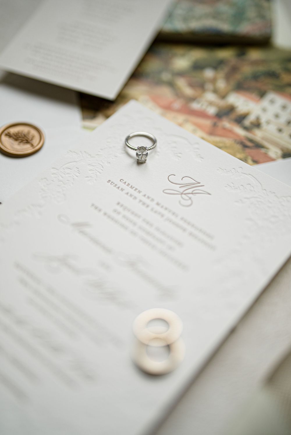An elegant diamond engagement ring rests on a white embossed wedding invitation with delicate monogram details.