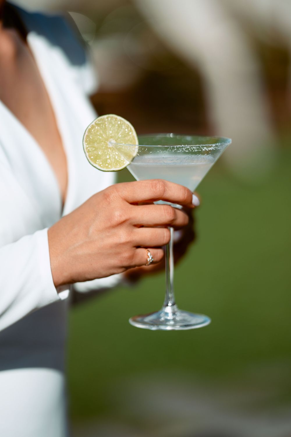 A martini glass with salt rim and lime garnish is held by a hand wearing a silver ring.