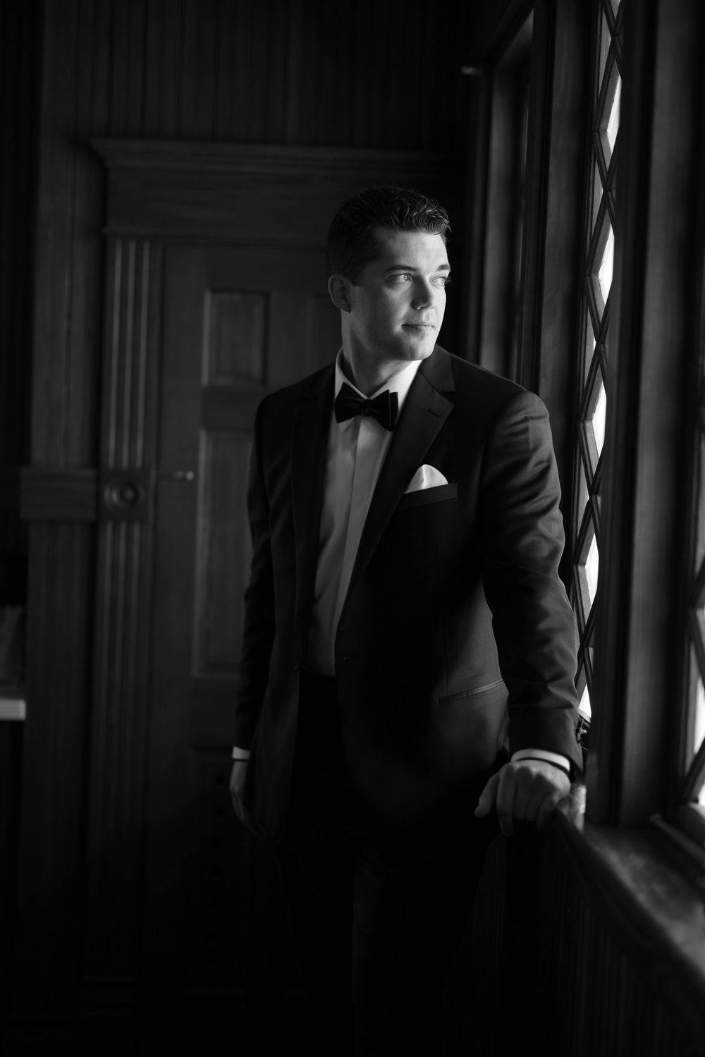Dramatic black and white portrait in formal attire showing person sitting by window with moody lighting and shadows.