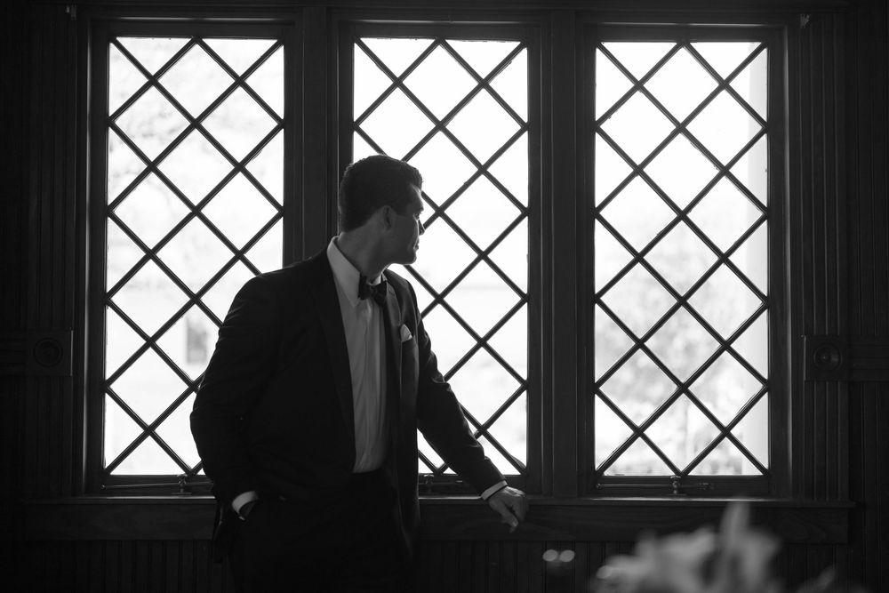 Silhouetted figure in formal attire stands near diamond-patterned window in dramatic black and white lighting.