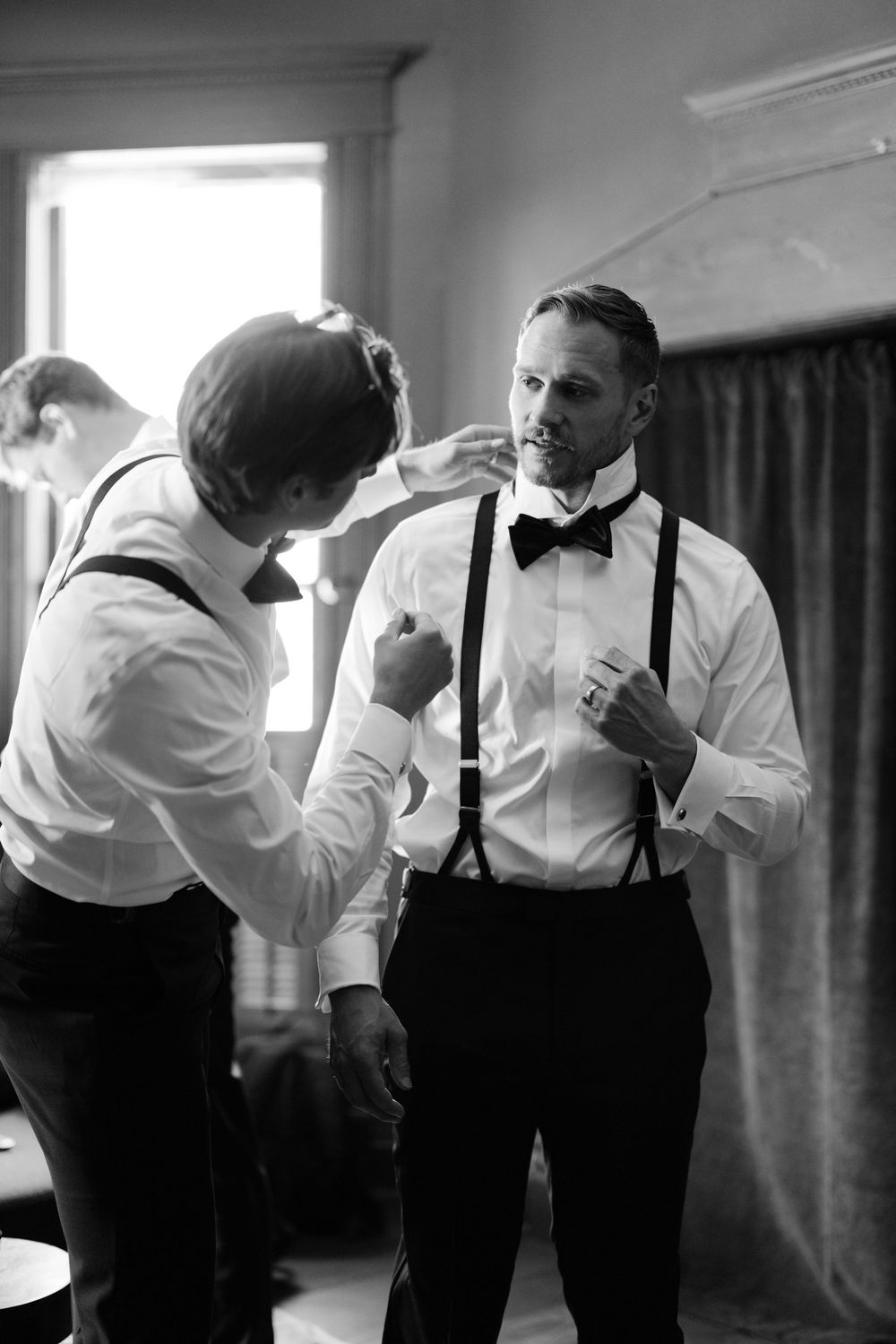 Two men in formal attire with bow ties and suspenders prepare for an event in black and white photography.