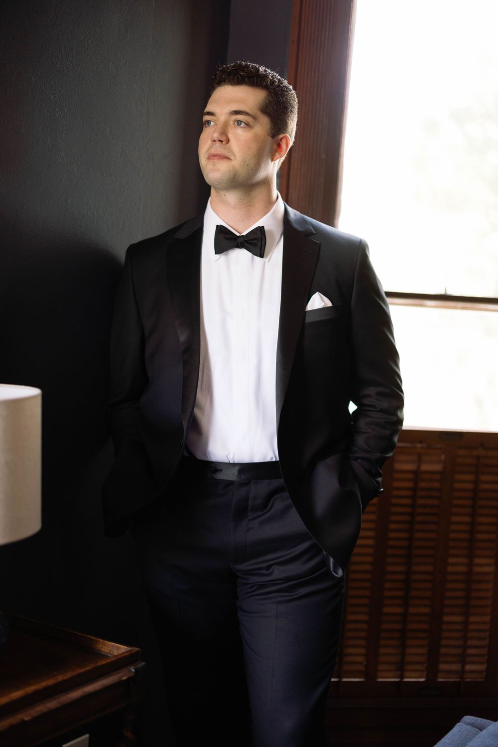 Elegant groom in classic black tuxedo poses thoughtfully by window in formal wedding photos series.