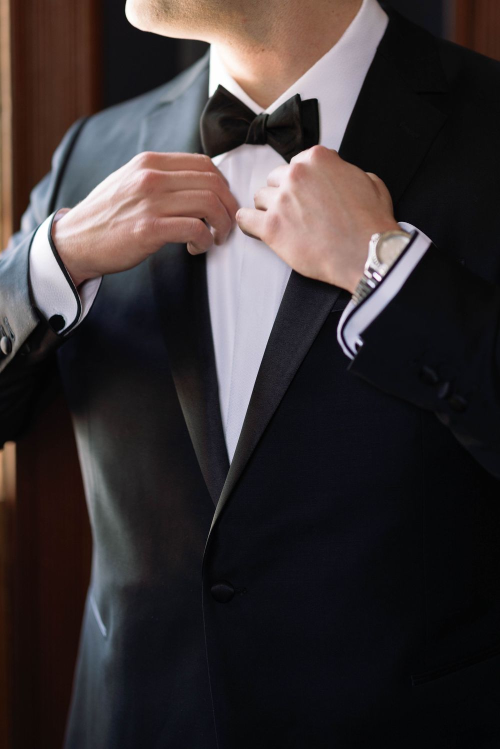 Close-up of hands adjusting black bow tie on classic tuxedo with white dress shirt and silver watch.