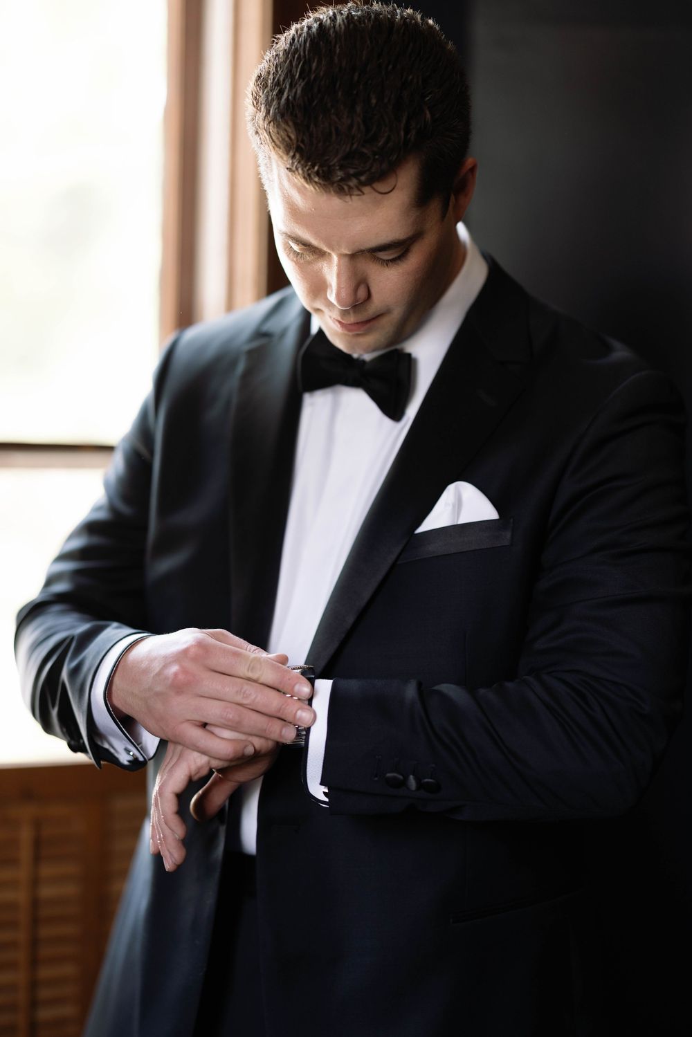 Groom in classic black tuxedo and bow tie adjusts his sleeve while getting ready on wedding day.