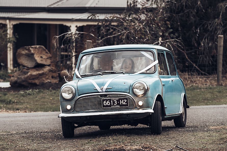 A classic light blue Austin Mini Cooper drives down a rural road with vintage styling from the early 1960s era.
