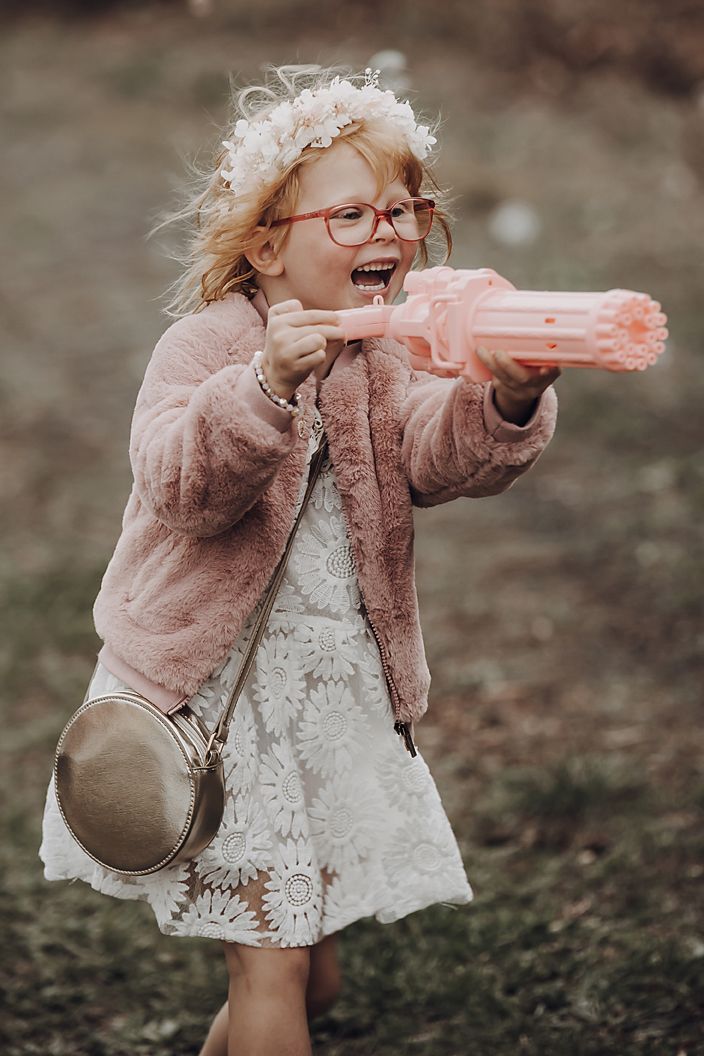 A child in a pink furry jacket and white dress joyfully plays with a pink toy outside.