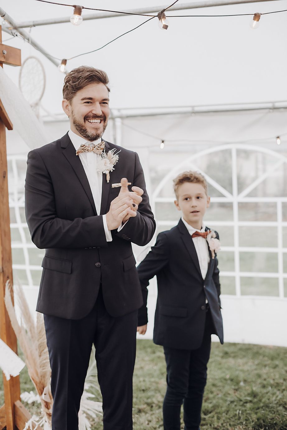 Two formally dressed groomsmen in black tuxedos and bow ties stand under twinkling lights at an outdoor wedding reception.