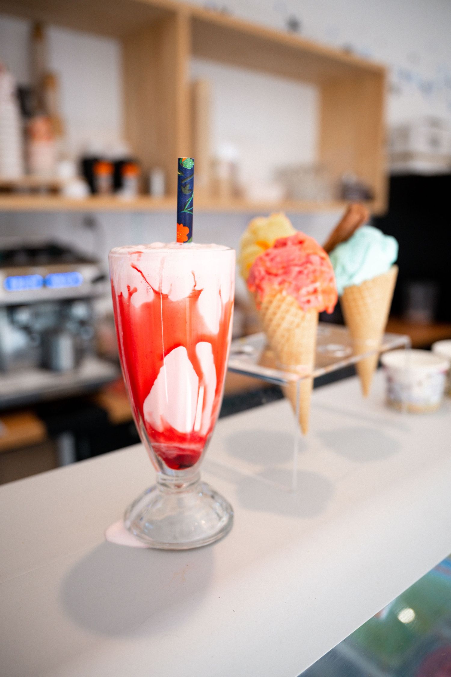 A tall milkshake glass filled with red syrup and ice cream, with ice cream cones displayed in the background.