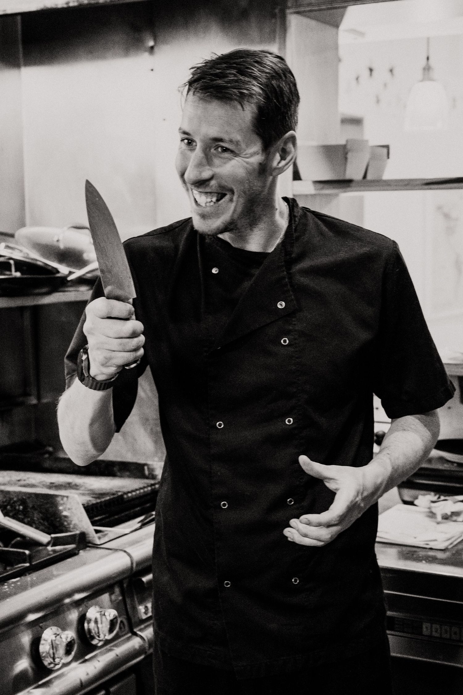 A chef in black uniform smiles while holding a kitchen knife in a commercial kitchen setting.