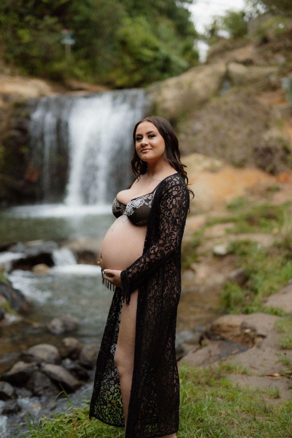 A maternity photoshoot featuring a figure in black lace against a serene waterfall backdrop in nature.