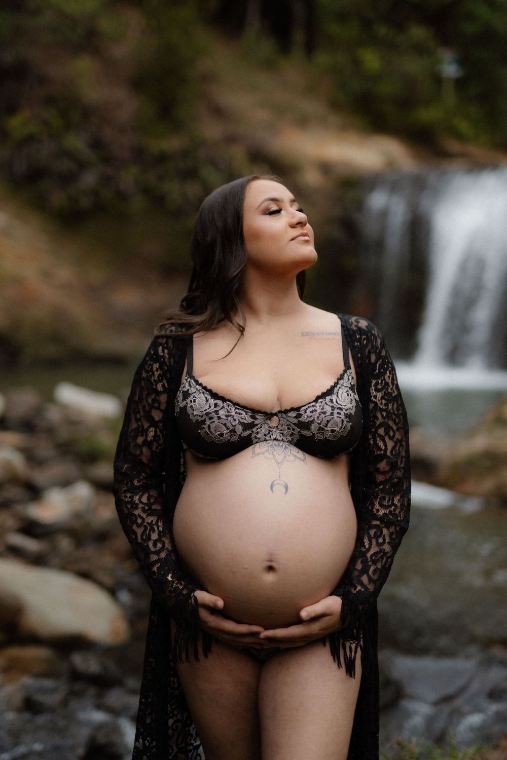 Maternity photos of an expectant mother in black lace dress posing near cascading waterfalls in nature.