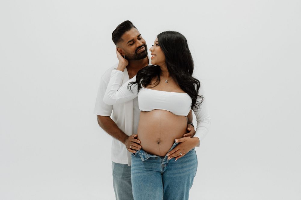A couple in casual attire share an intimate moment against a white background during a maternity photoshoot.