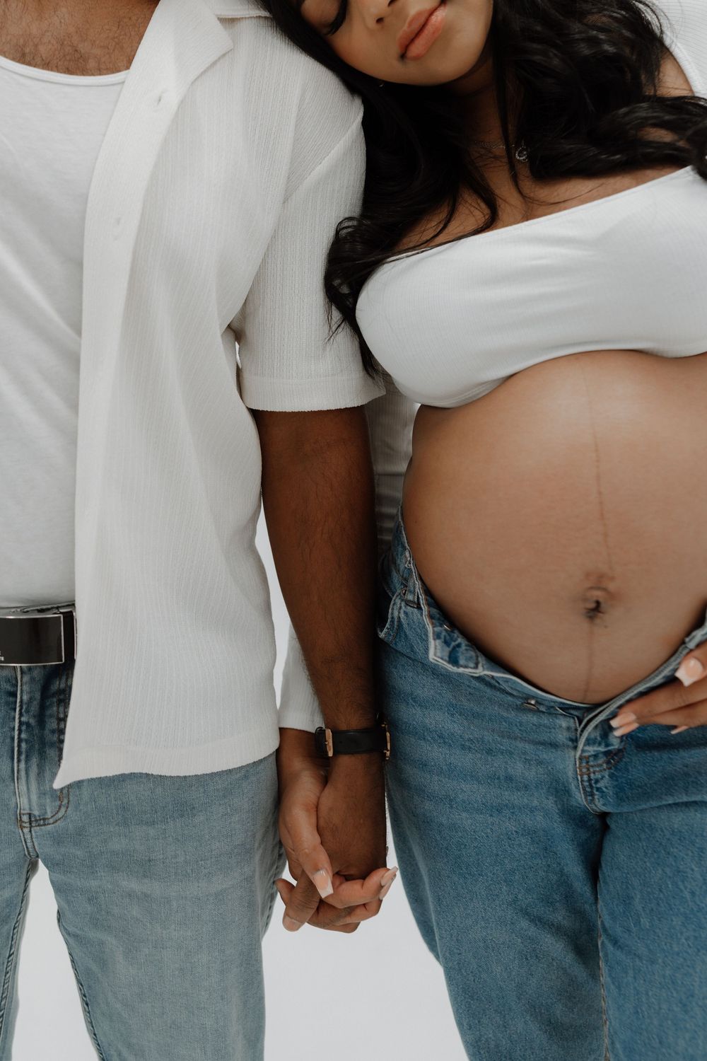 Intimate close-up of couple holding hands in casual white tops and jeans, with woman showing bare pregnant belly.