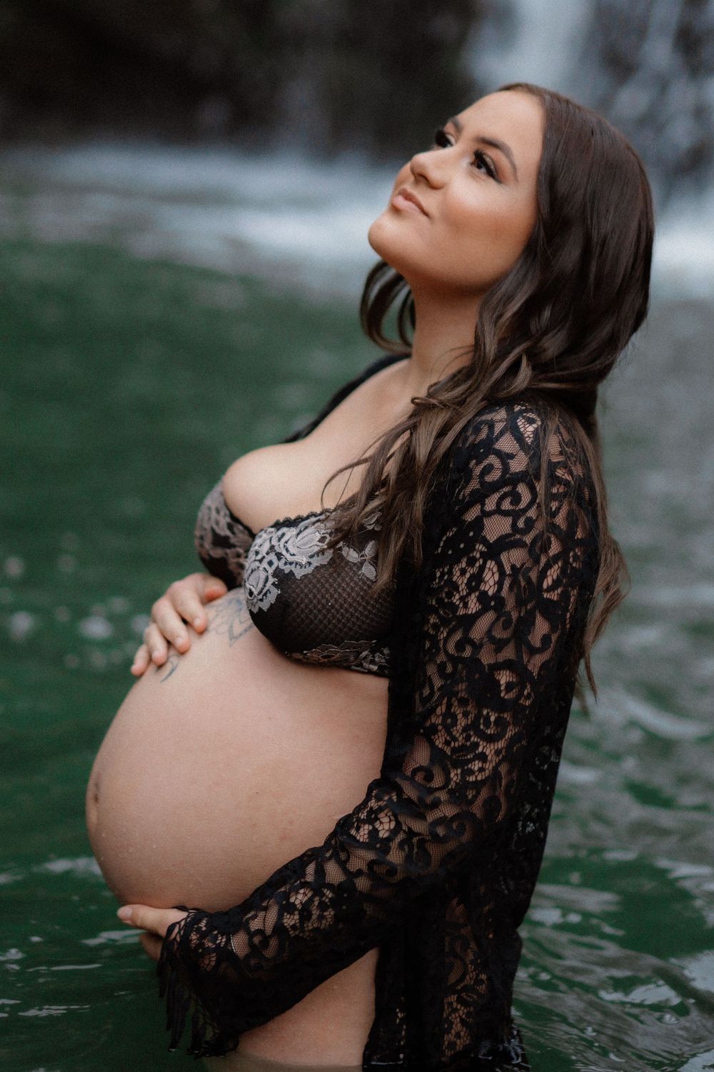 Beautiful maternity portrait of someone in black lace and sequins standing in emerald green water during sunset.