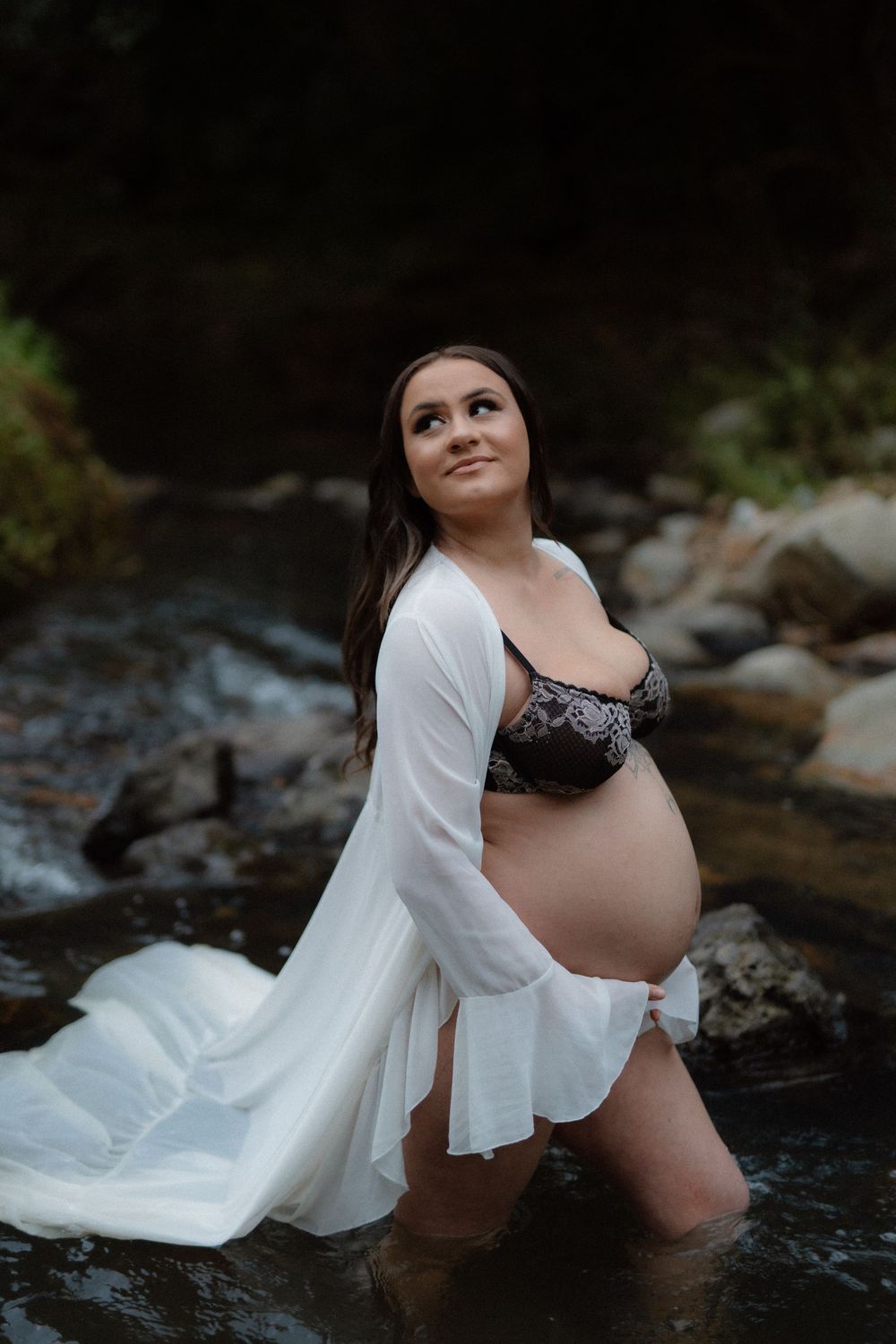 Maternity photo shoot in a stream with figure wearing white flowing robe in natural outdoor setting.