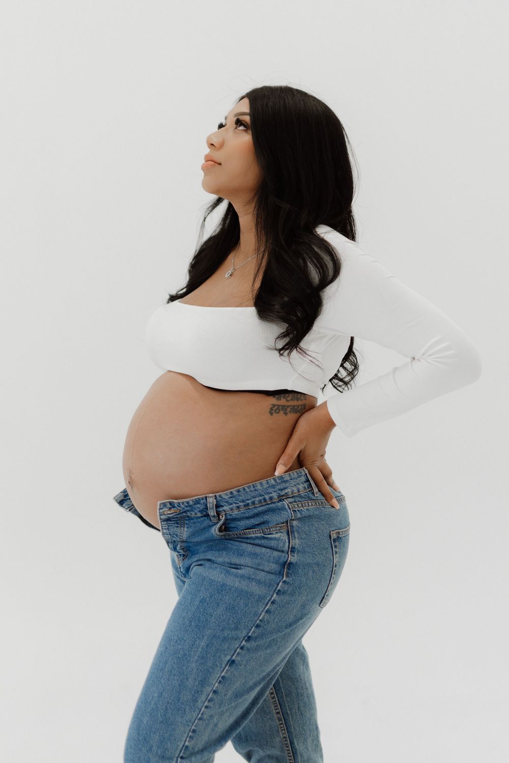 Side view of a pregnant woman in a white crop top and blue jeans posing against a white background.