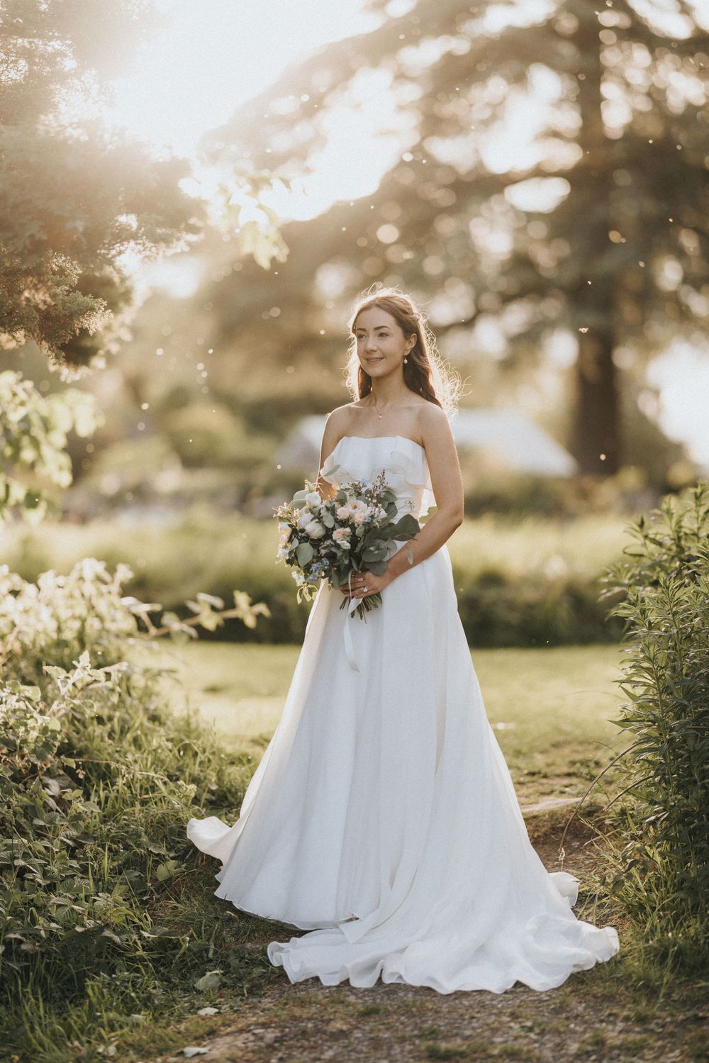 A flowing white wedding dress is captured in golden sunlight against a dreamy outdoor garden backdrop.