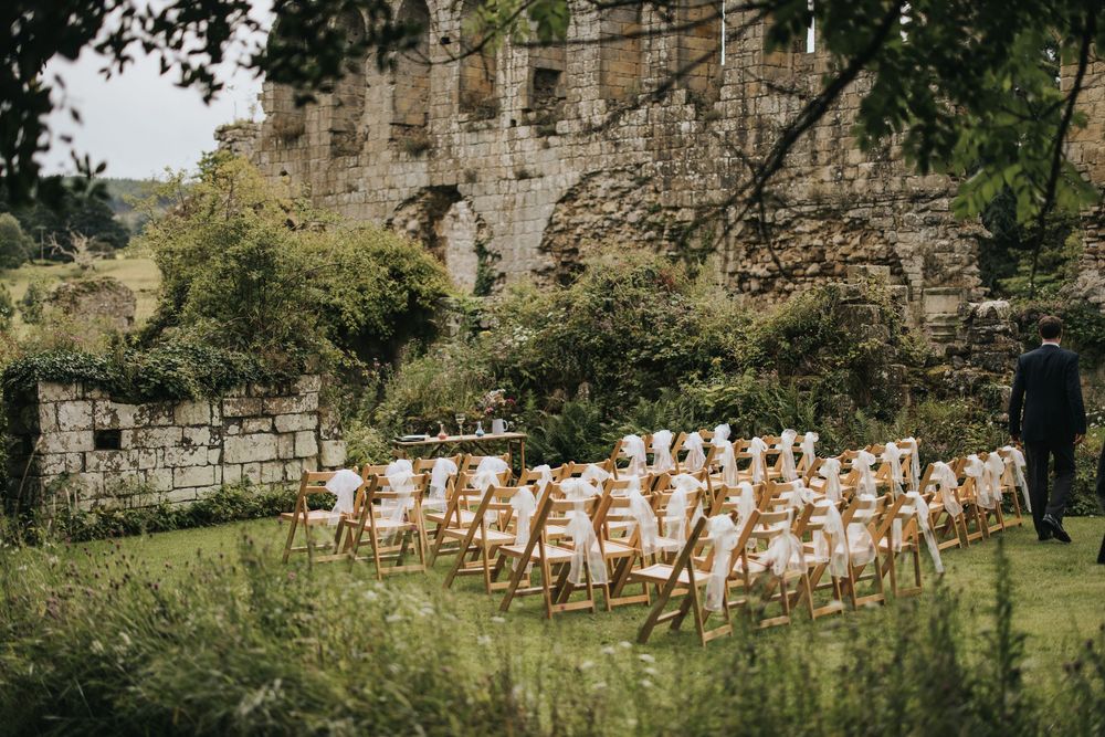 White wooden chairs arranged for an outdoor wedding ceremony against ancient stone castle ruins.