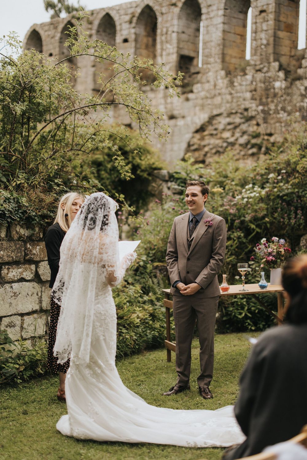 Outdoor wedding ceremony in front of stone ruins with gothic arches and greenery on a sunny day.