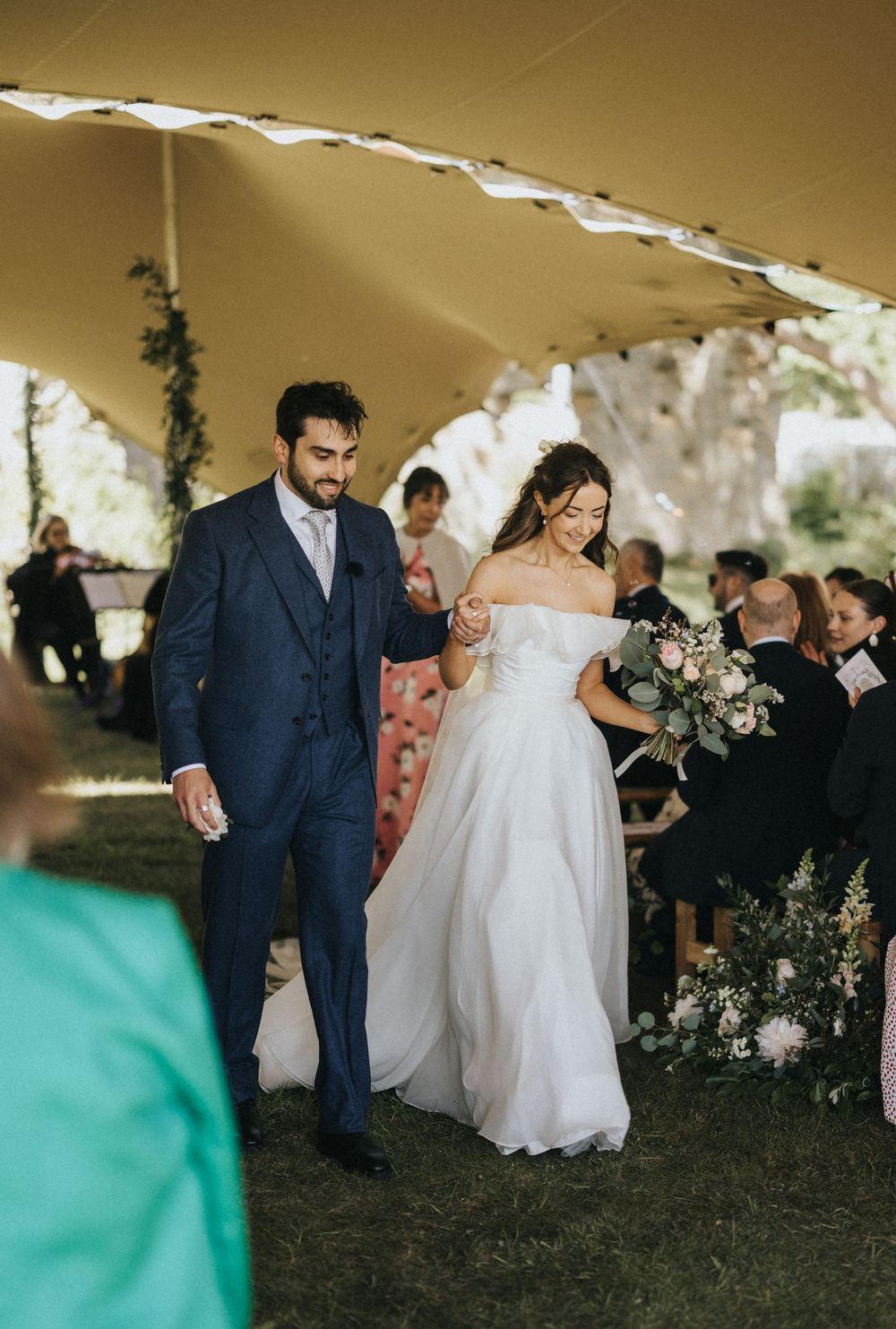 A wedding couple walks down the aisle under a tent canopy while guests look on during an outdoor ceremony.