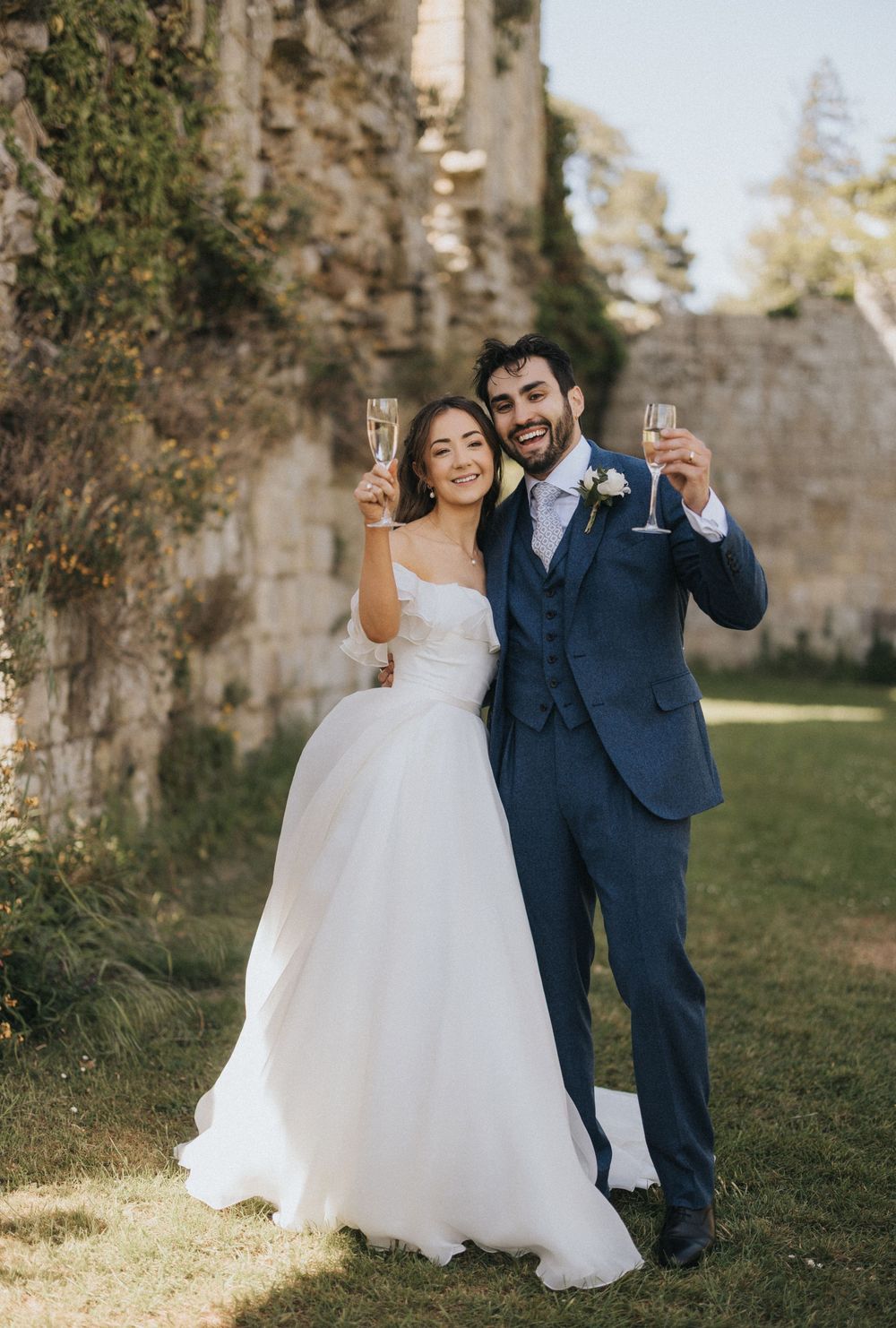 A happy couple toasts champagne in their wedding attire against a rustic stone wall backdrop outdoors.