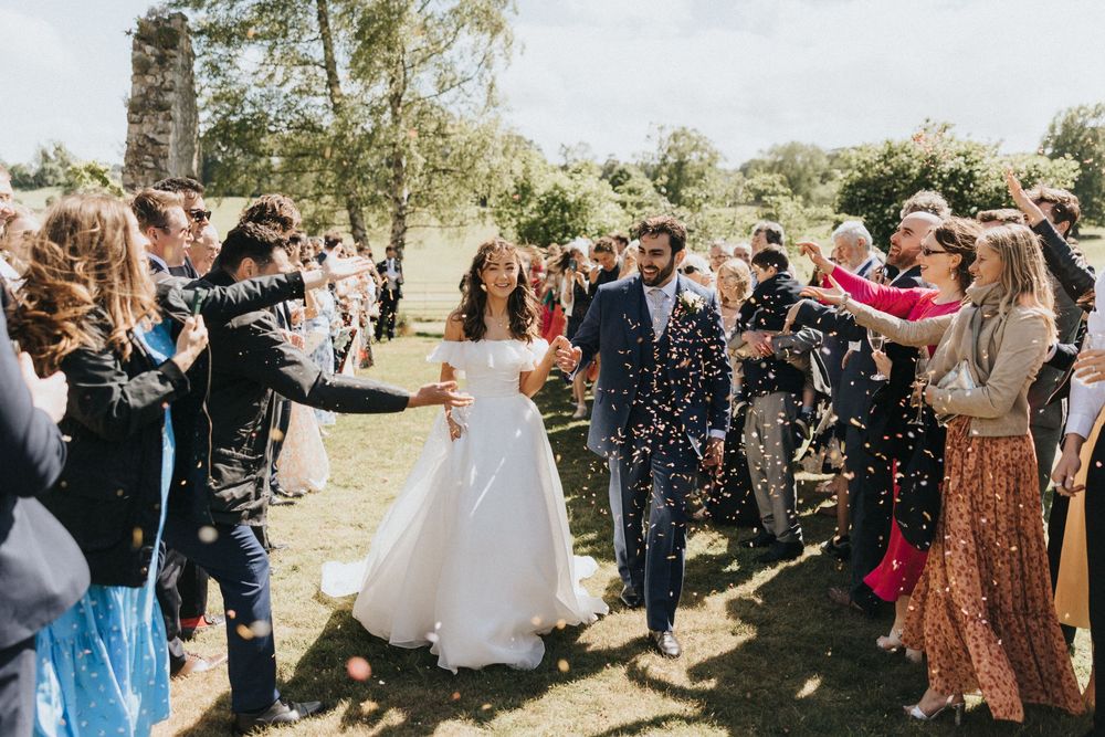 Wedding guests toss confetti and celebrate during an outdoor ceremony in a sunlit garden setting.