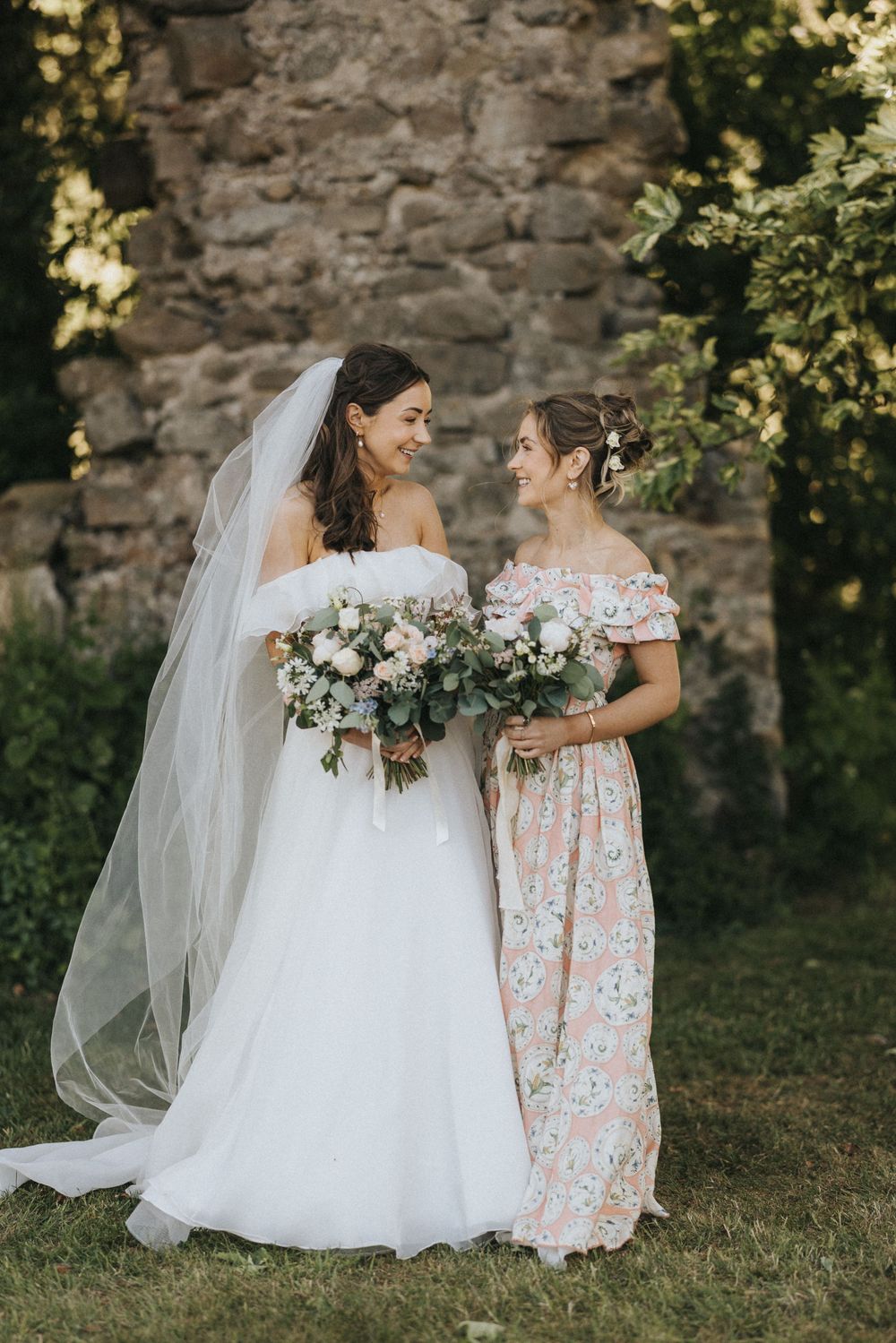 Two people stand together outdoors wearing off-shoulder dresses, one in white with a veil holding bouquets against stone wall.