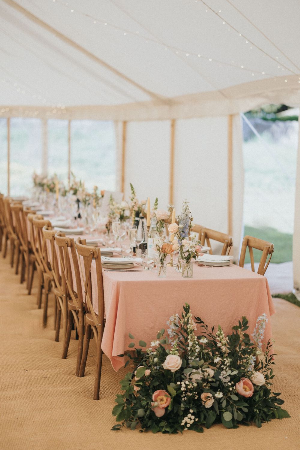Elegant wedding reception tables set with dusty pink linens, wooden chairs, and lush floral centerpieces in a light-filled tent.