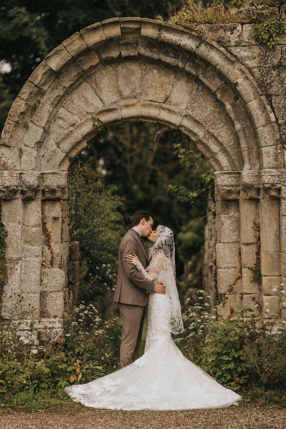 A romantic wedding photo beneath a historic stone archway surrounded by greenery and foliage.