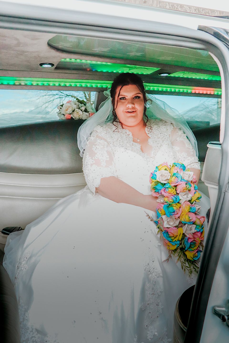 A bride in a white lace wedding dress sits in a vehicle decorated with green LED lights while holding a colorful bouquet.