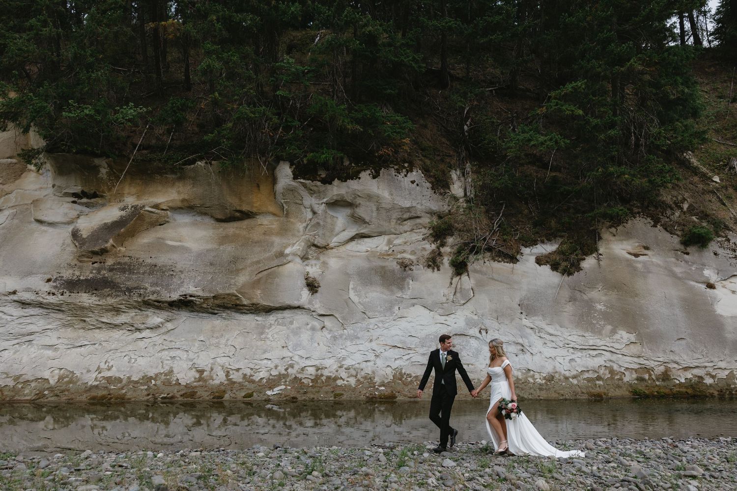 A couple in wedding attire walks near a rocky cliff at sunset in a dramatic natural landscape.