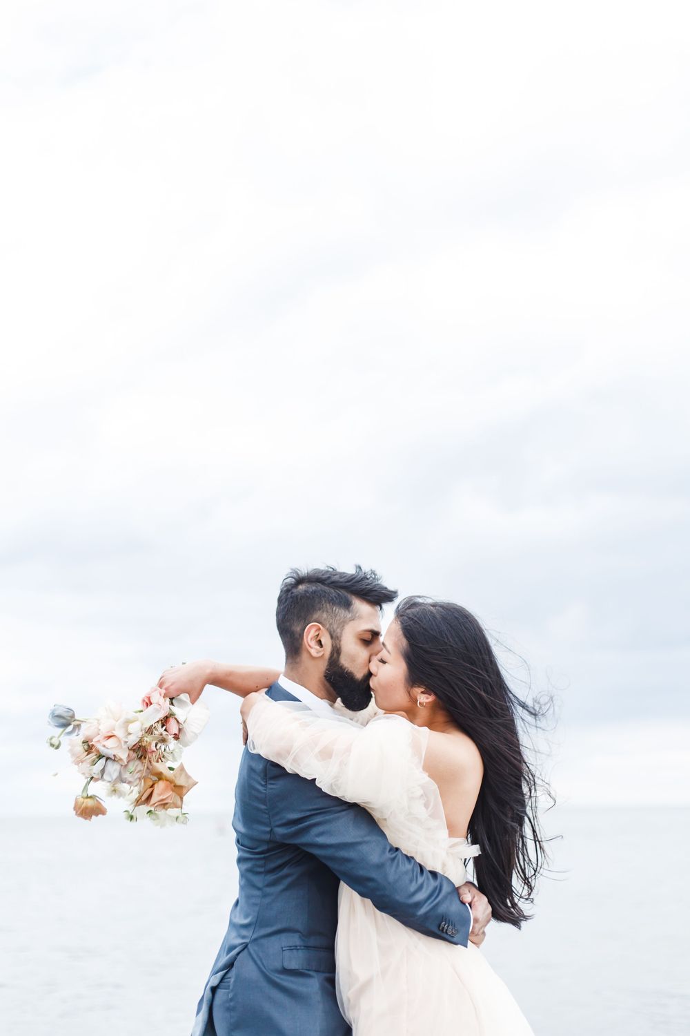 Romantic couple in wedding attire dancing and twirling on a beach under cloudy skies.