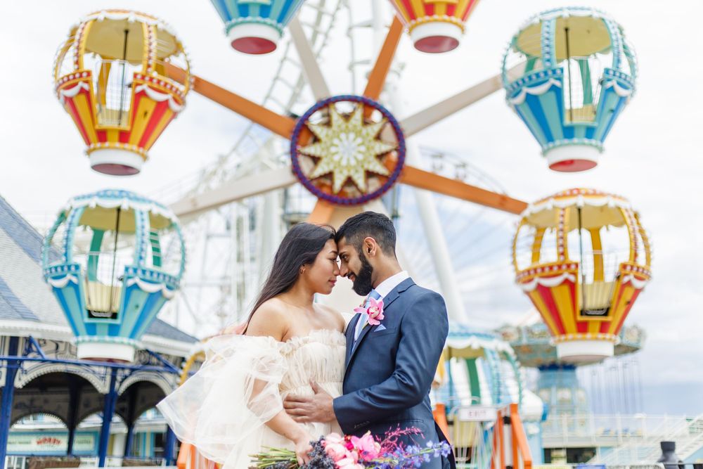 Newlyweds sharing a romantic moment in front of a colorful carnival ferris wheel with decorative gondolas.