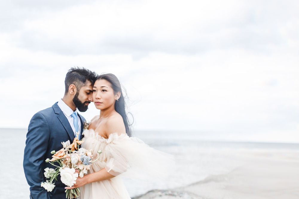 Newlyweds embracing on a cloudy beach with bride holding a white floral bouquet.