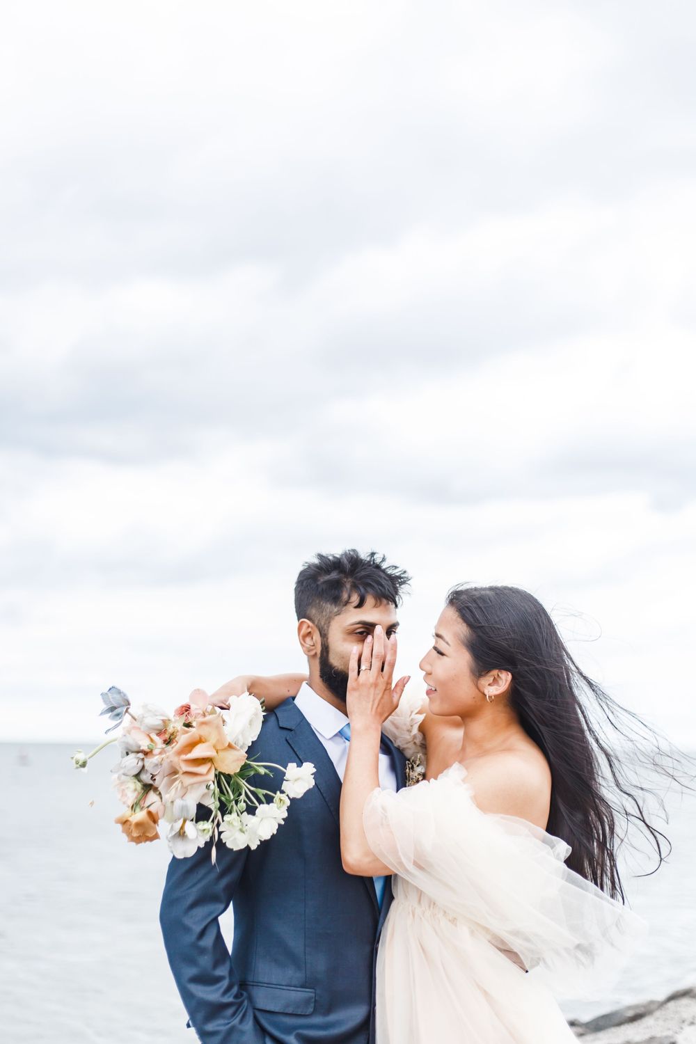 A couple shares a playful romantic moment on a cloudy beach while holding a floral bouquet.