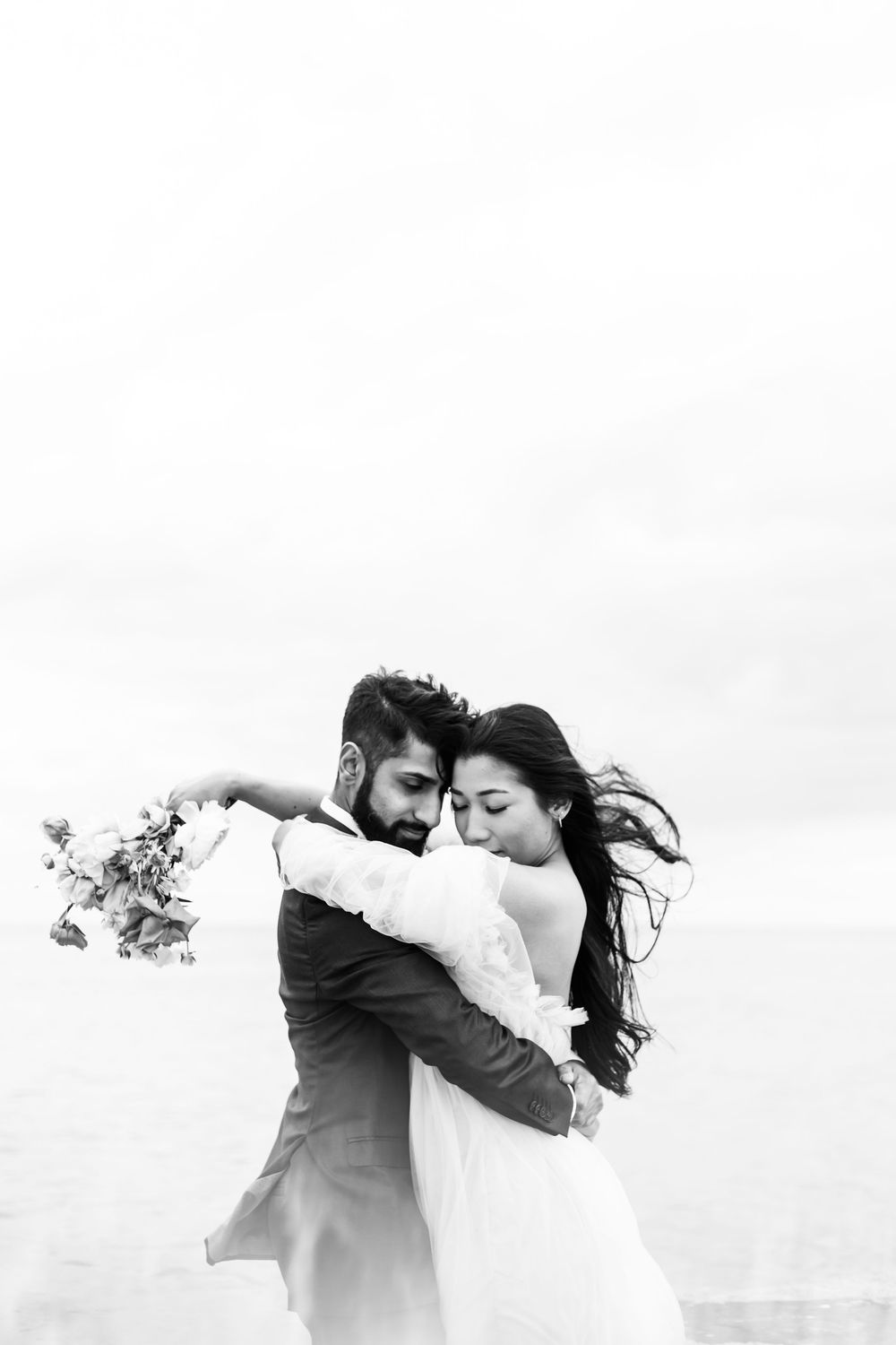 A couple in wedding attire share a romantic embrace against a bright sky.