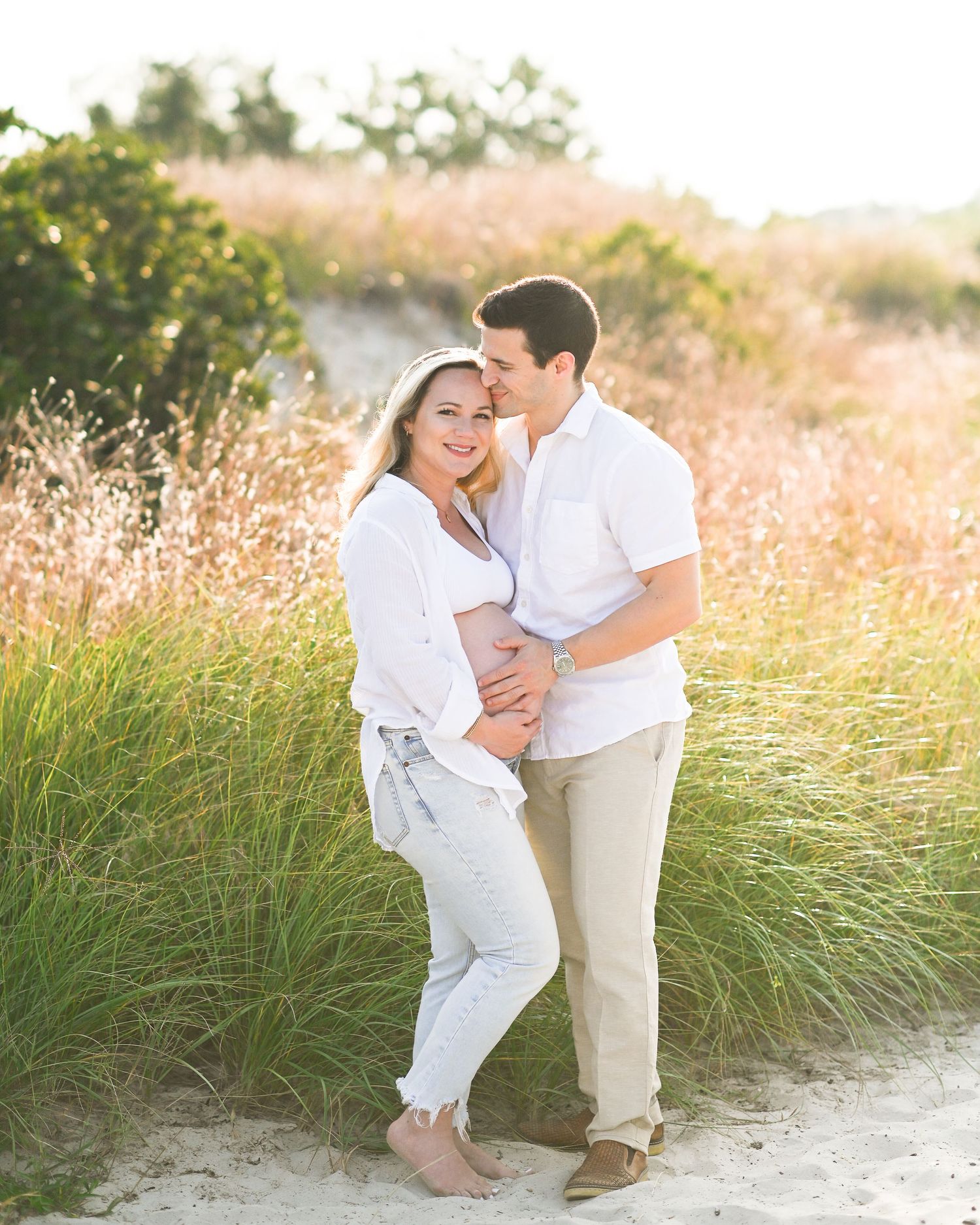 A romantic couple in white outfits embrace on a sunlit path surrounded by golden sea grass during sunset.