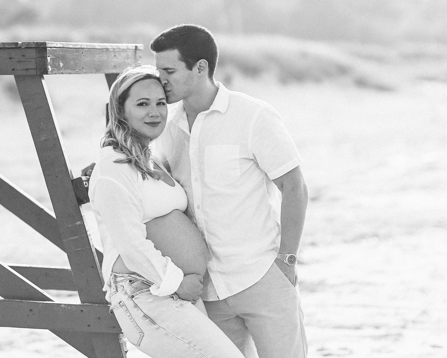 A romantic black and white portrait of a couple embracing against a wooden fence on a beach.