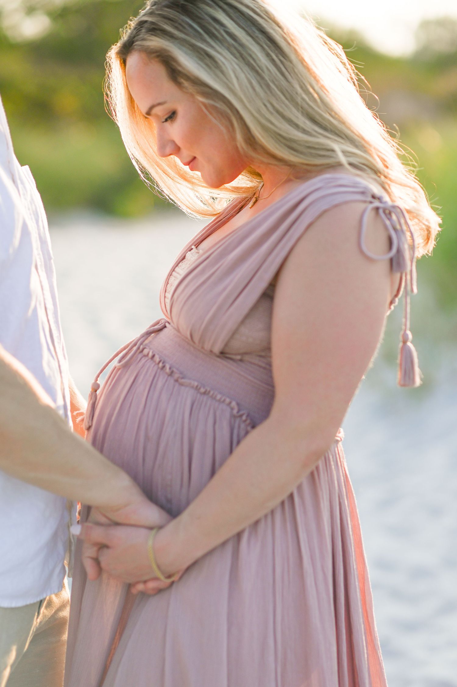 A glowing maternity photo features an expectant mother in a flowing blush pink dress during a golden sunset.