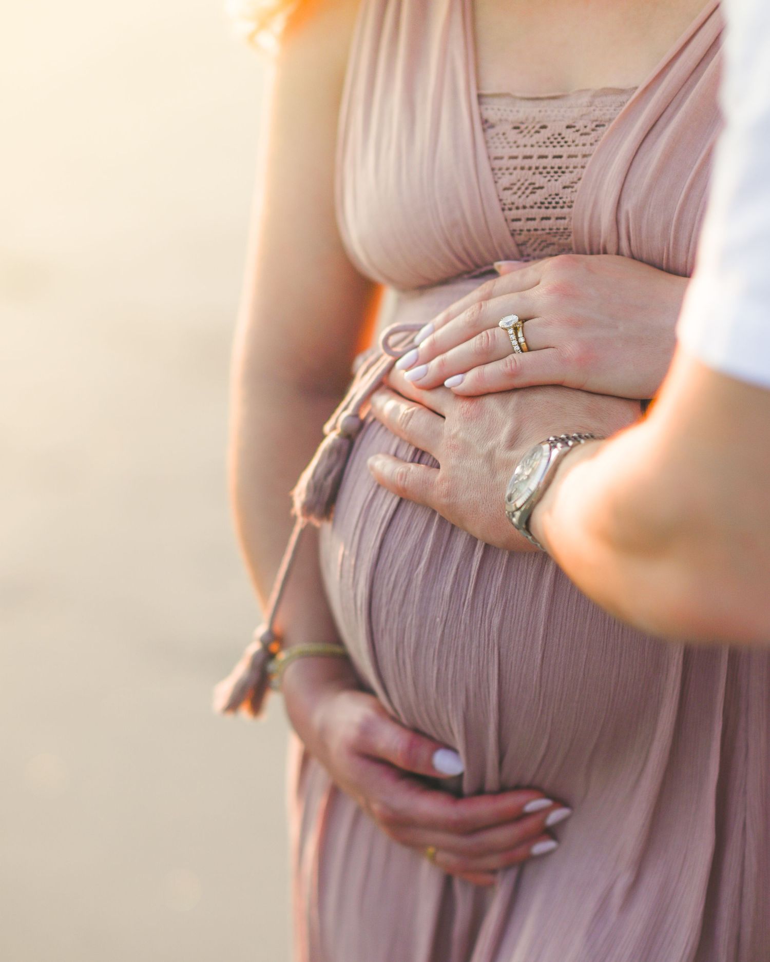 Close up view of pregnant belly in flowing mauve dress with hands elegantly placed showing rings and jewelry.
