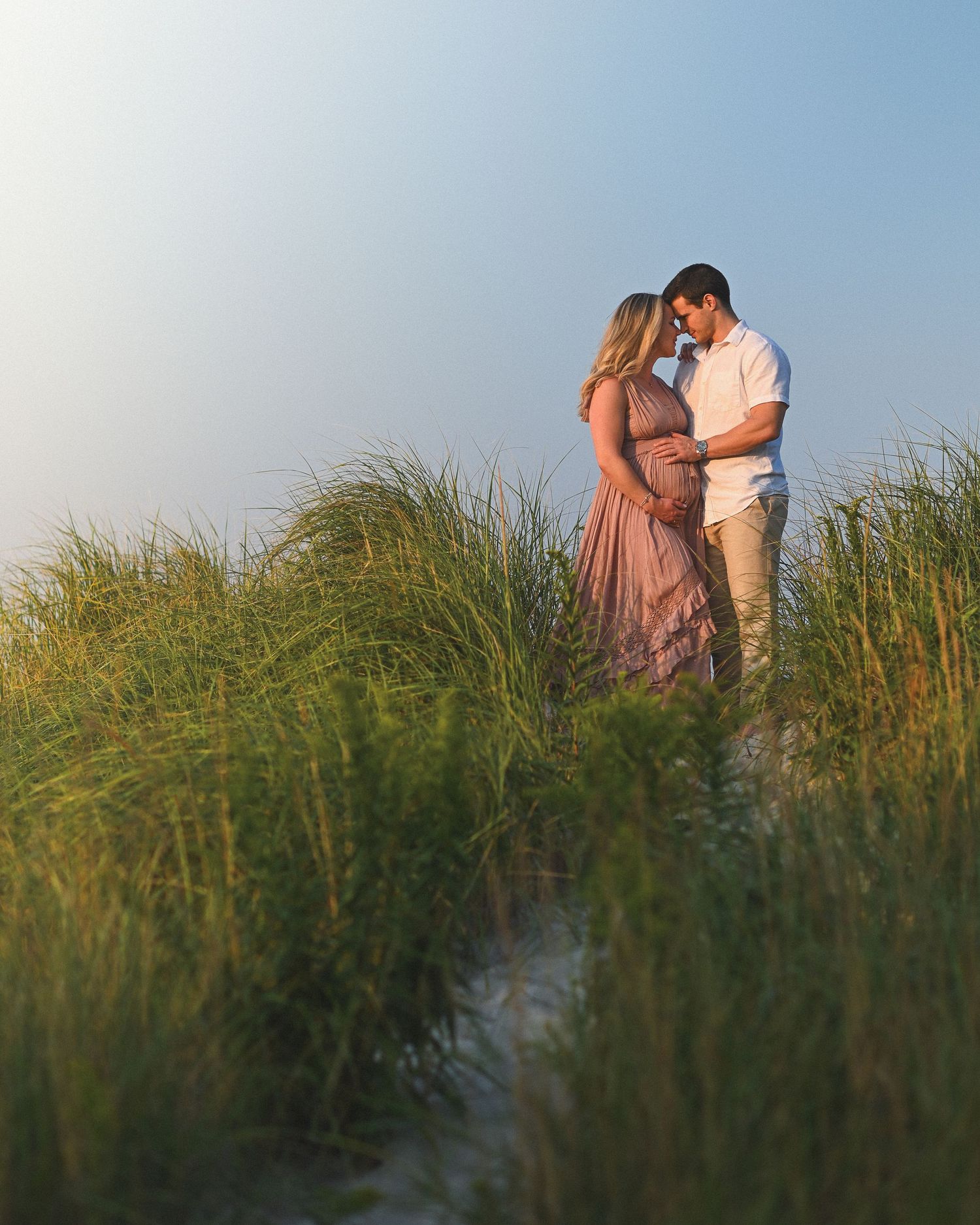 A couple embraces on a beach at sunset among tall grass dunes creating a romantic silhouette.