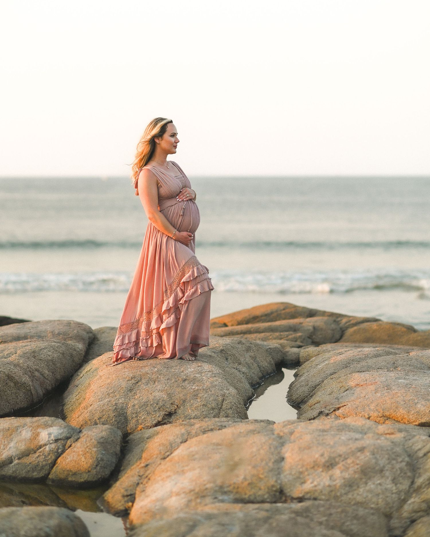 A person in a flowing peach dress sits peacefully on coastal rocks at sunset overlooking the ocean.