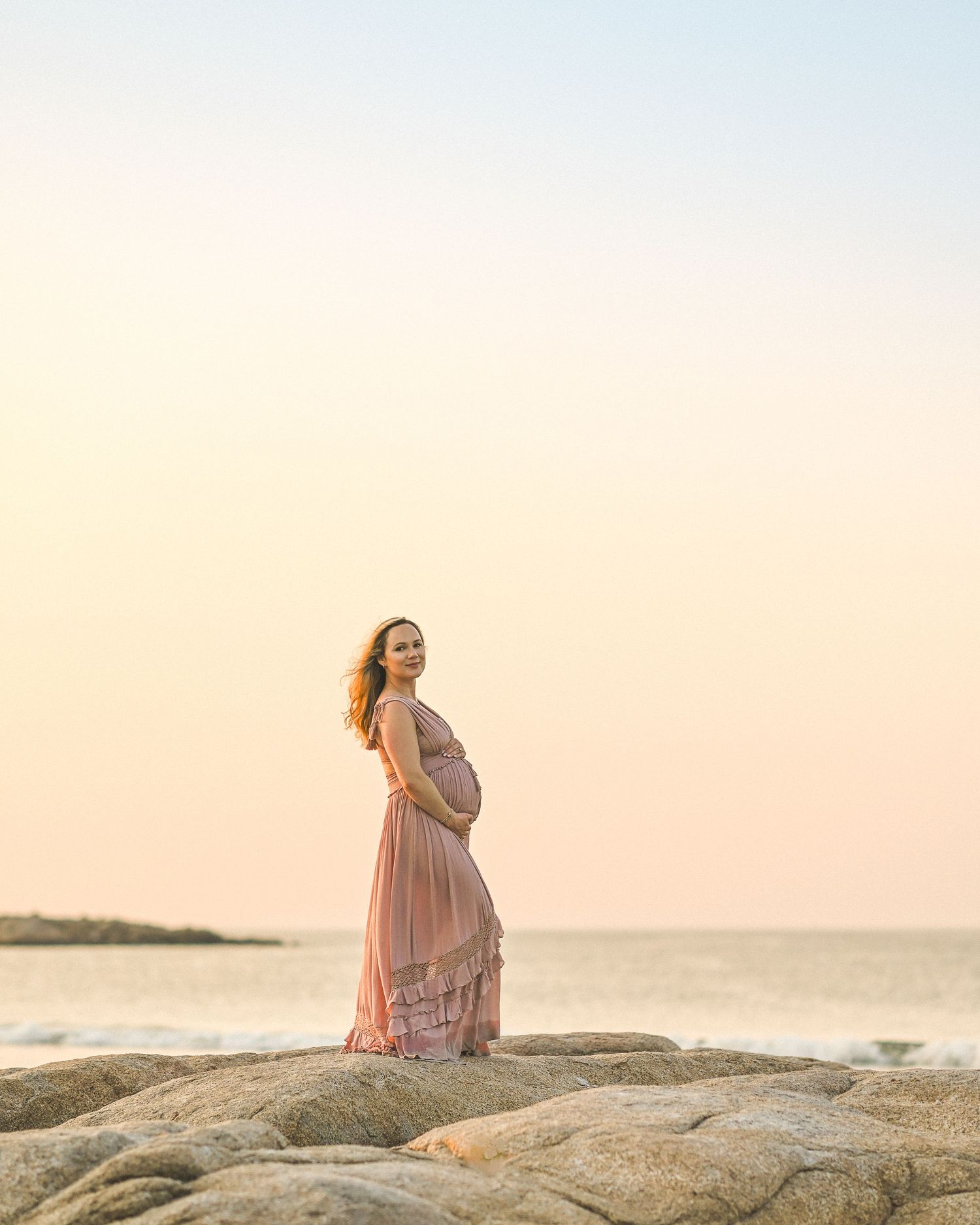 Silhouette of pregnant woman in flowing pink dress standing on coastal rocks at sunset.