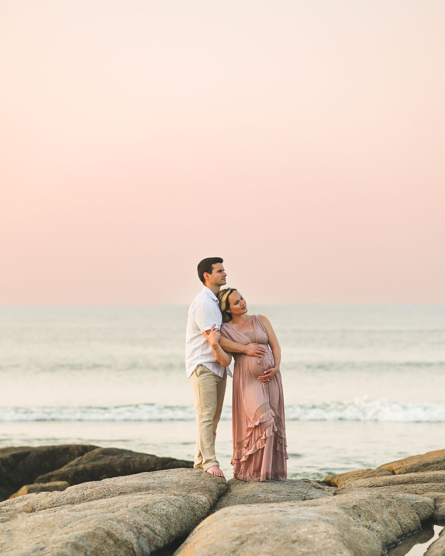 Two people sitting together on rocky coastal outcrops against a soft pink sunset sky and ocean backdrop.