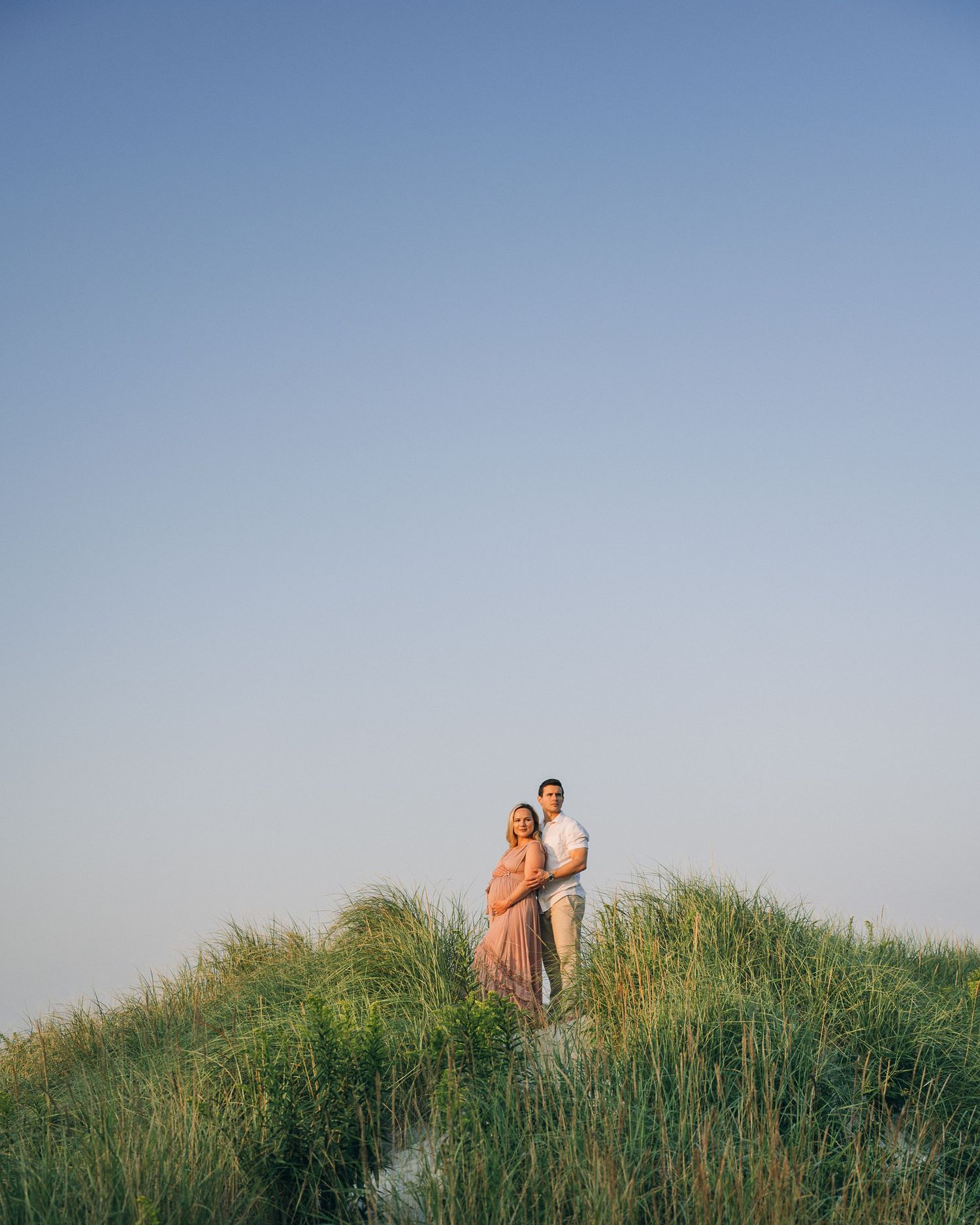 A couple stands together on a grassy coastal dune against a clear blue sky.